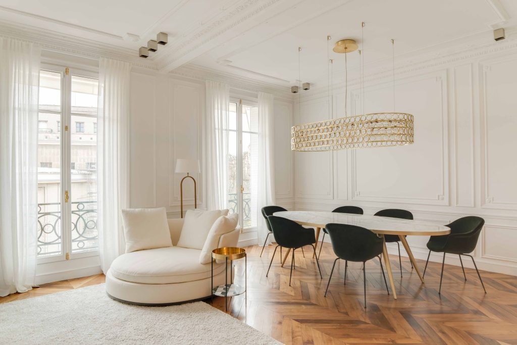 Sala em estilo clássico, com paredes, cortinas e sofá branco. Um lustre oval dourado. Mesa redonda branca com detalhes dourados e cadeiras pretas