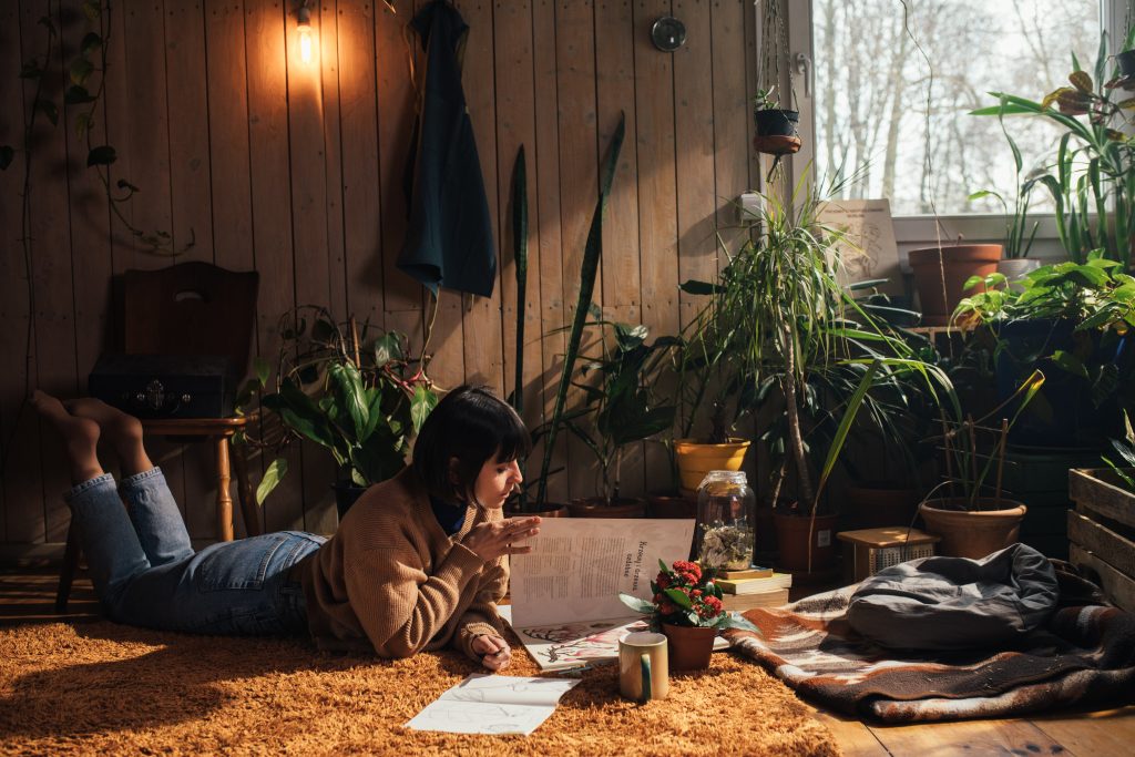 Tendências de arte e design: bem-estar. Uma mulher deitada no chão, de barriga para baixo, lendo uma revista. ao lado dela, uma xícara e um cobertor. Ao redor dela, muitas plantas.