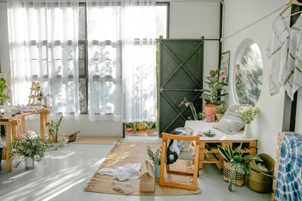 Sala de paredes e cortinas brancas, tapete bege e mesas de madeira. Porta na cor verde e muitas plantas.